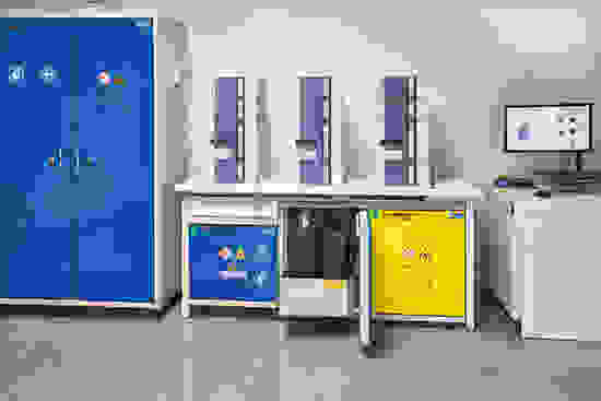 Laboratory workspace featuring three stacked storage units, a computer setup, and two cabinets with safety warning labels, one yellow and one blue.