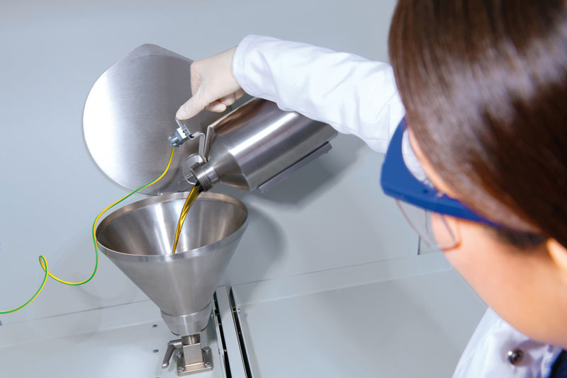 A laboratory technician in a white coat pours liquid from a stainless steel container into a funnel.