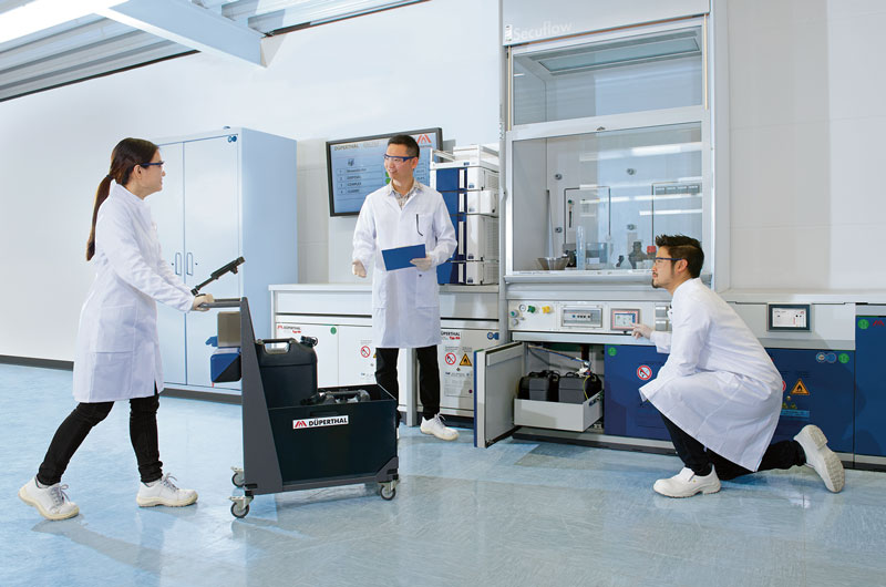 Three people in lab coats work in a laboratory setting, using a cart with equipment and interacting with lab equipment and controls.