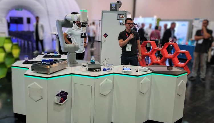 Ein Messestand mit einem Pr&auml;sentierenden, einem kollaborativen Roboter und einem modernen, hexagonalen Tisch.