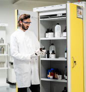 Un technicien de laboratoire portant une blouse blanche tient une bouteille dans une chambre de stockage de produits chimiques contenant plusieurs r&eacute;cipients.