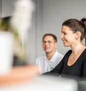 Une femme souriante est assise devant un ordinateur, tandis qu'un homme regarde en arri&egrave;re-plan.
