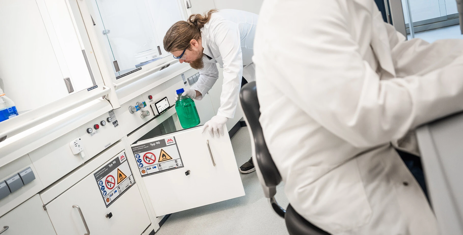 Une personne en blouse de laboratoire et portant des gants saisit un liquide vert dans un laboratoire. À l'arrière-plan, des équipements de laboratoire et des panneaux de sécurité sont visibles.