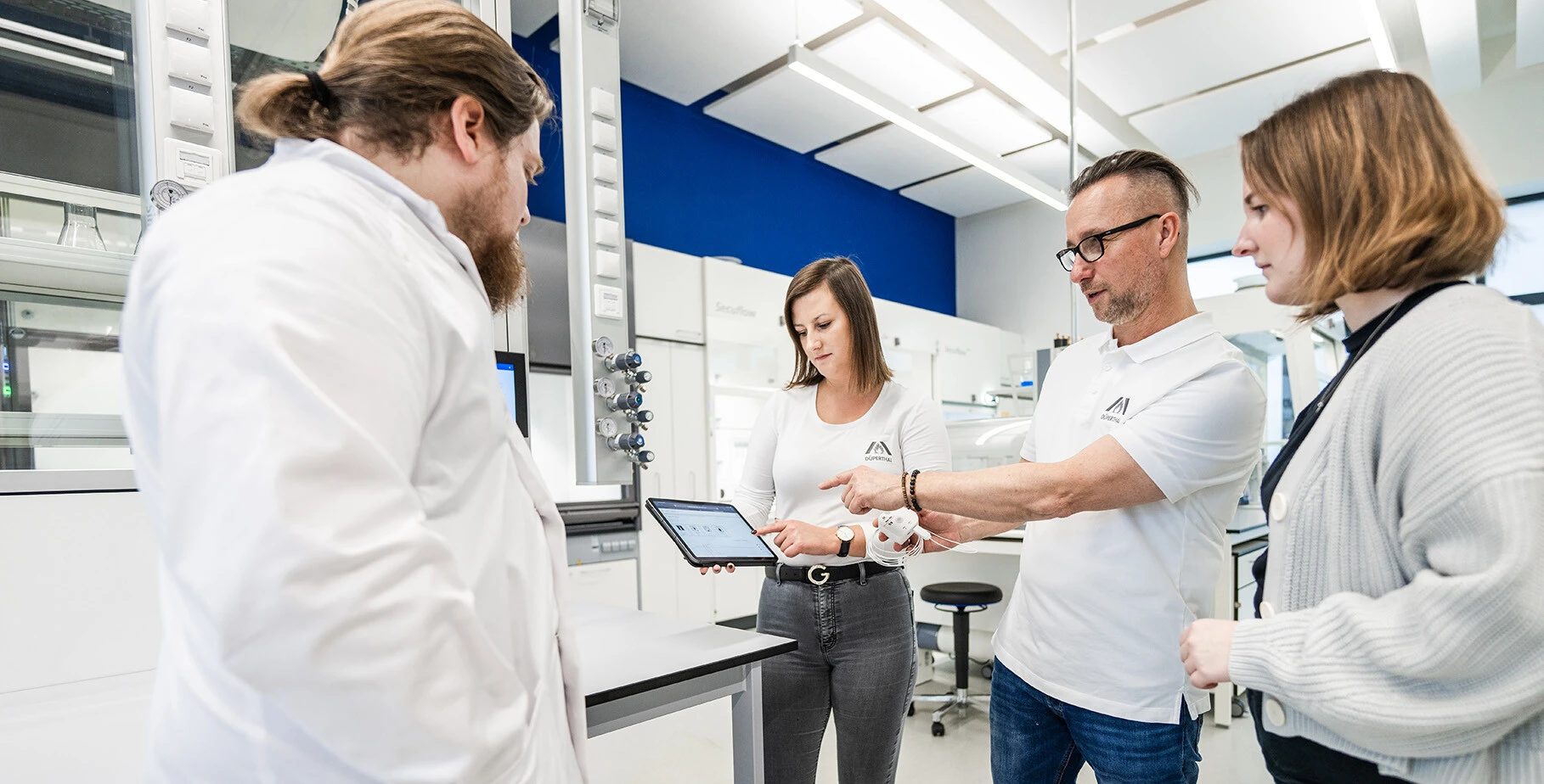 Quatre personnes dans un laboratoire regardent une tablette. Ils discutent, tandis que des appareils et du matériel de laboratoire sont visibles en arrière-plan.