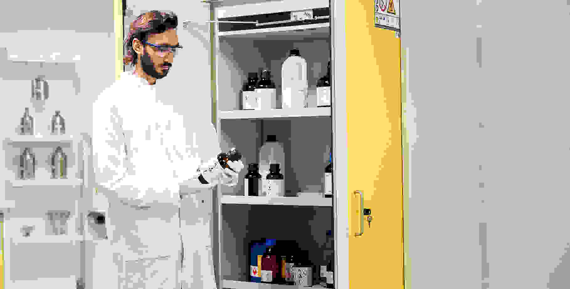 Un technicien de laboratoire en blouse blanche se tient devant une armoire remplie de bouteilles de produits chimiques et tient dans la main une bouteille de substance dangereuse.