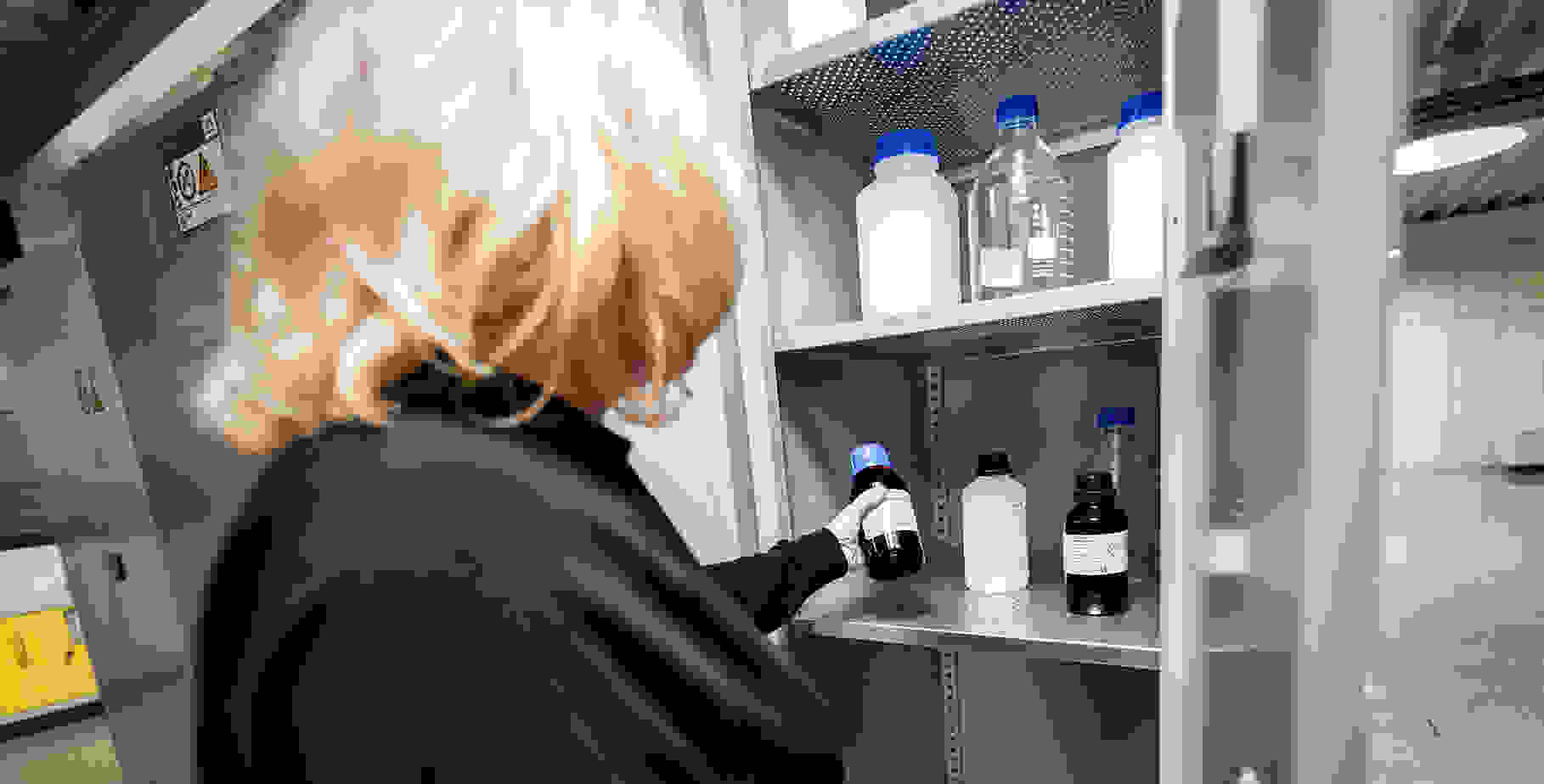 Une personne aux cheveux blonds se tient devant une armoire contenant des bouteilles et des récipients chimiques.