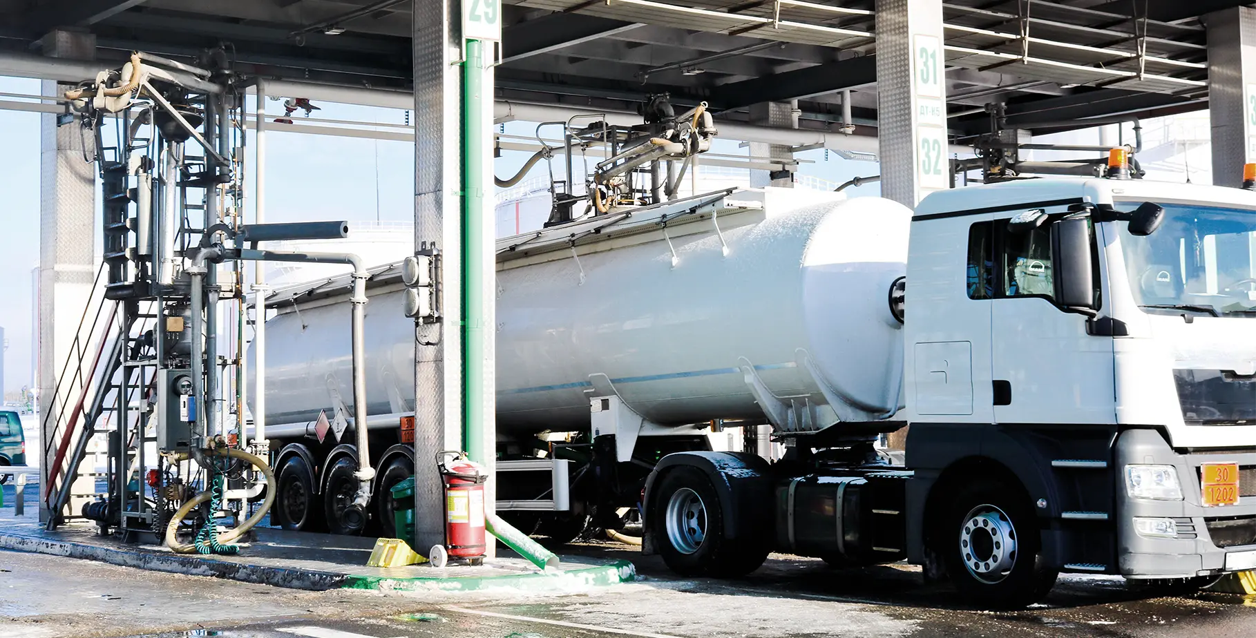 Un camion avec un grand réservoir dans une station-service, entouré de tuyauteries et de pompes.