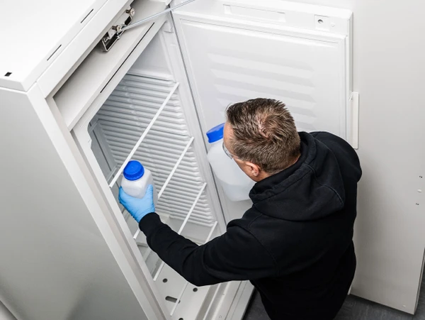 Un homme portant des gants tient un récipient dans la porte vide d'un réfrigérateur.