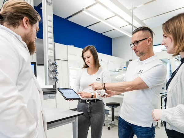 Quatre personnes dans un laboratoire regardent une tablette. Ils discutent, tandis que des appareils et du matériel de laboratoire sont visibles en arrière-plan.