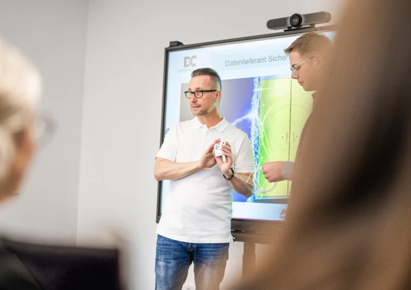 Deux hommes présentent devant un écran dans une salle de réunion. L'homme à gauche montre un appareil, tandis que l'autre fait office d'auditeur. L'écran montre des graphiques.