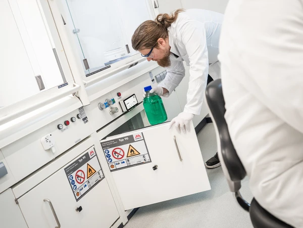 Une personne en blouse de laboratoire et portant des gants saisit un liquide vert dans un laboratoire. À l'arrière-plan, des équipements de laboratoire et des panneaux de sécurité sont visibles.